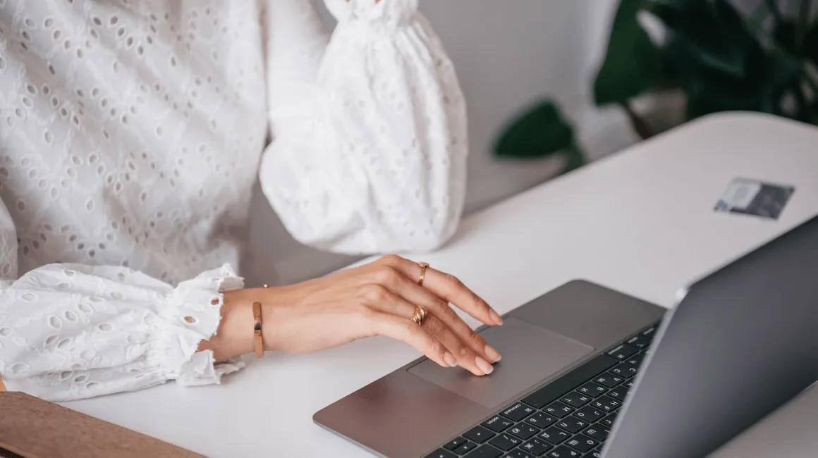 Kobieta w białej bluzce obsługuje laptopa, widoczne jej dłonie i biżuteria